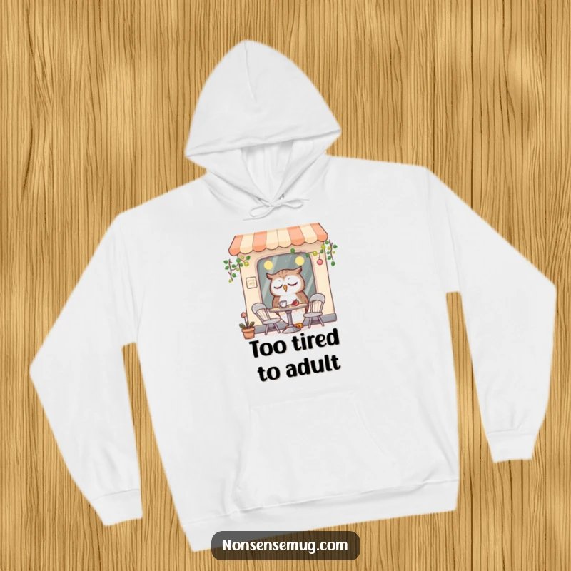 Funny hoodie showcasing a drowsy owl character seated in a quaint, miniature cafe, offering comfort and a laugh.
